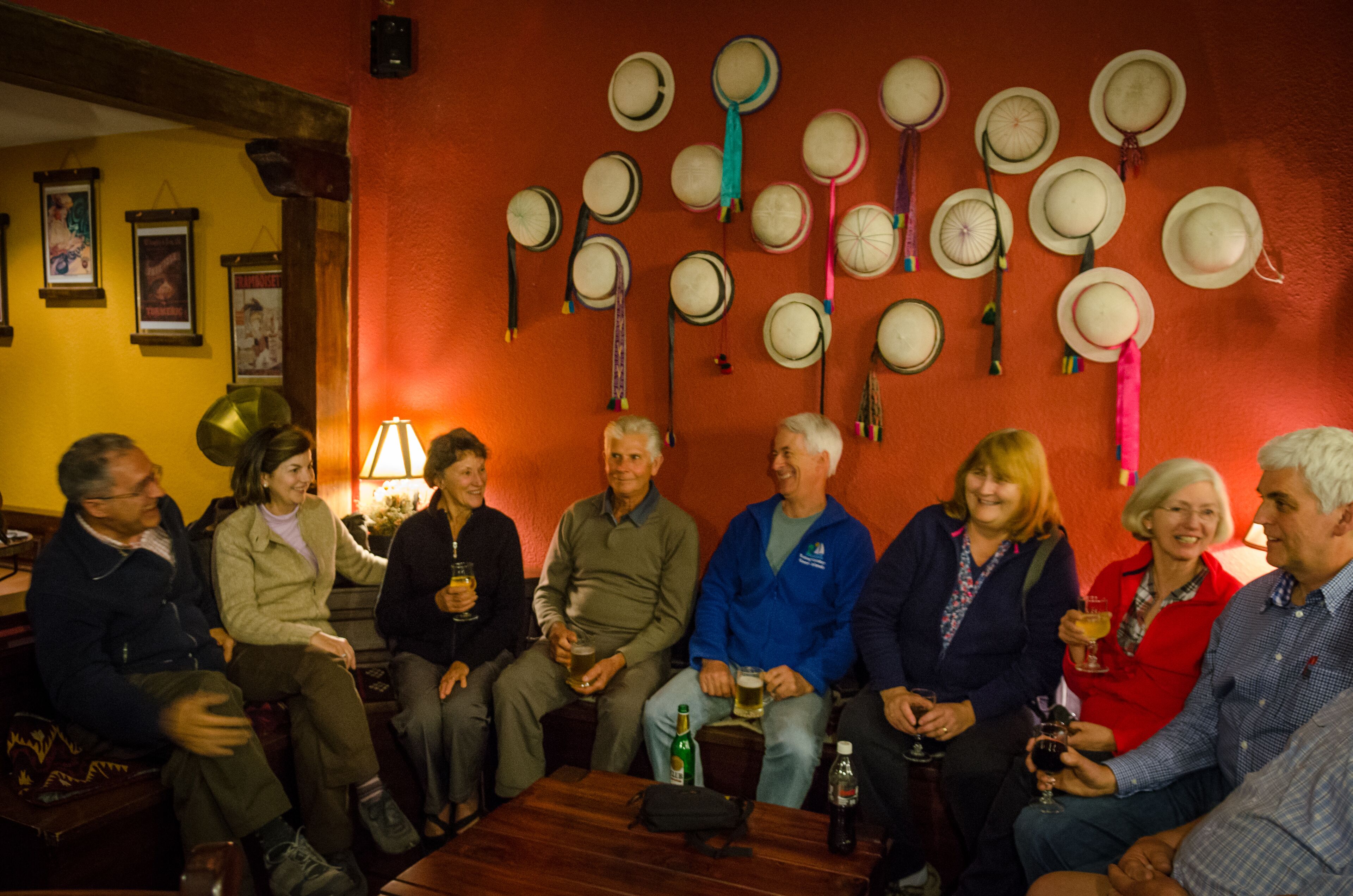 Un grupo de personas sentadas alrededor de una mesa en una habitación con pared roja, algunas sosteniendo bebidas.