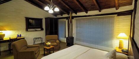 Un dormitorio con cama, dos sillones, una mesita, televisor y ventana con persianas.