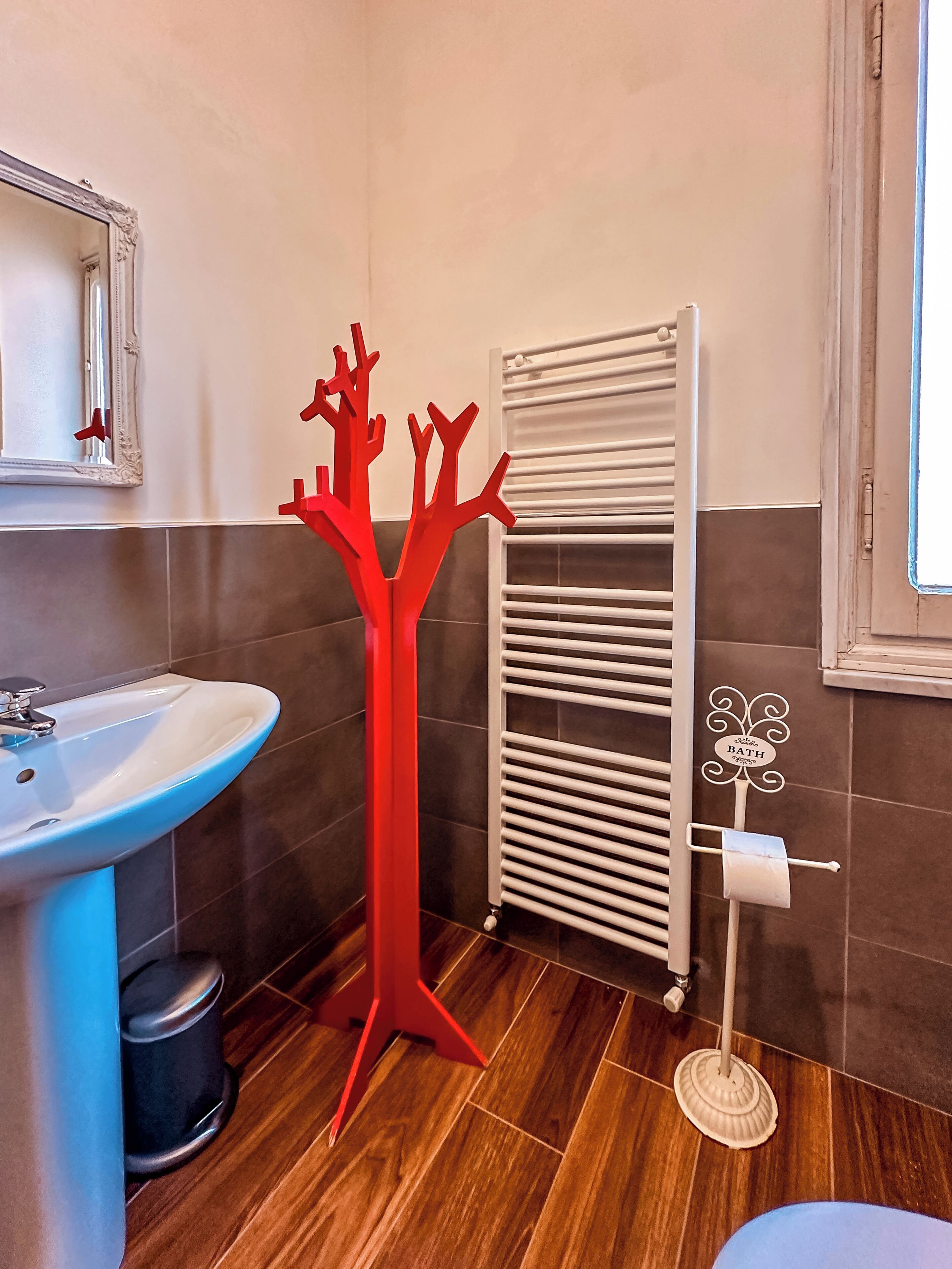 Ein Badezimmer mit einem weißen Waschbecken, einem roten abstrakten Baum-Dekorationsobjekt, einem weißen Heizkörper und einem Toilettenpapierhalter mit einem Schild.