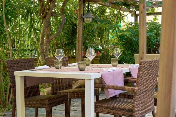 An outdoor dining area with wicker chairs and a white table, surrounded by lush greenery.