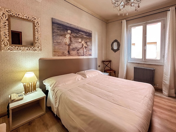 Un dormitorio con una cama grande, una mesita de noche con lámpara, una silla, un cuadro, un espejo y una ventana con cortinas.
