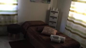 Une chambre à coucher avec un lit, un petit tabouret, une table de chevet, un tableau accroché au mur et des rideaux.