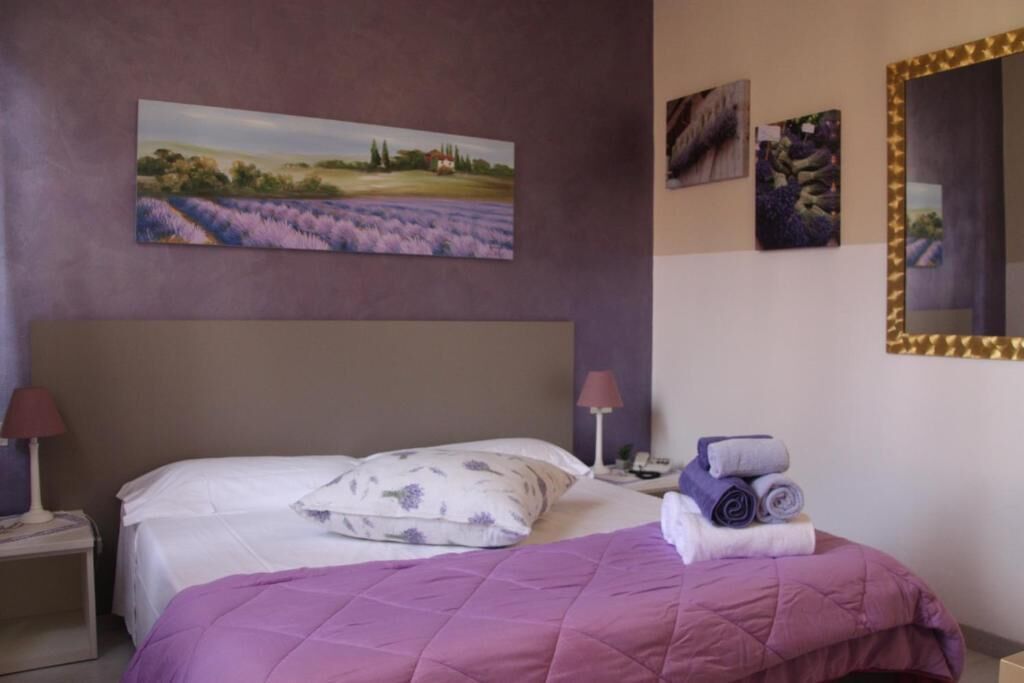 A neatly made bed with a purple quilt, a painting of lavender fields on the wall, and a mirror with a gold frame.