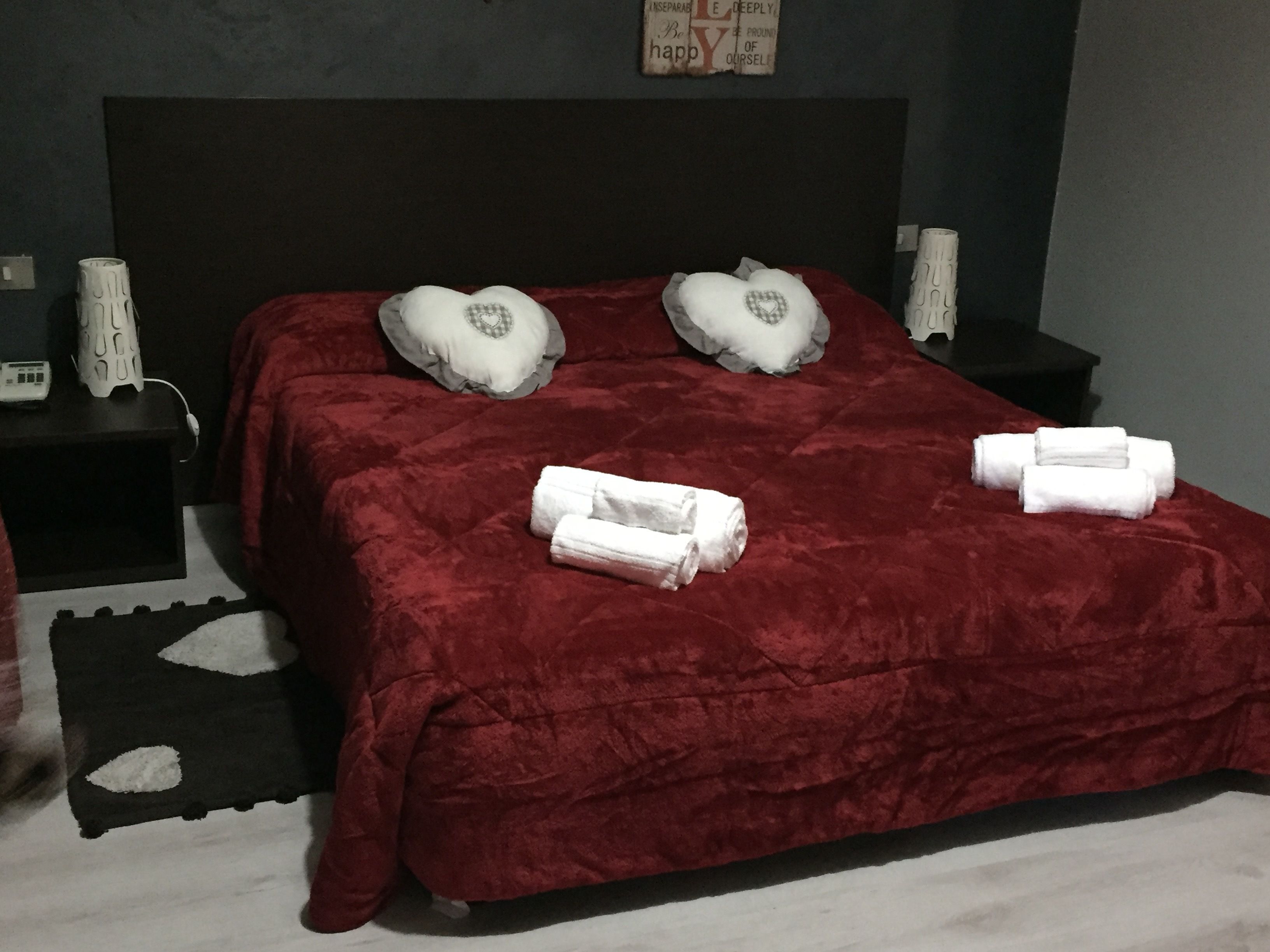 A neatly made bed with a red blanket, two heart-shaped pillows, and two rolled towels on the bed.