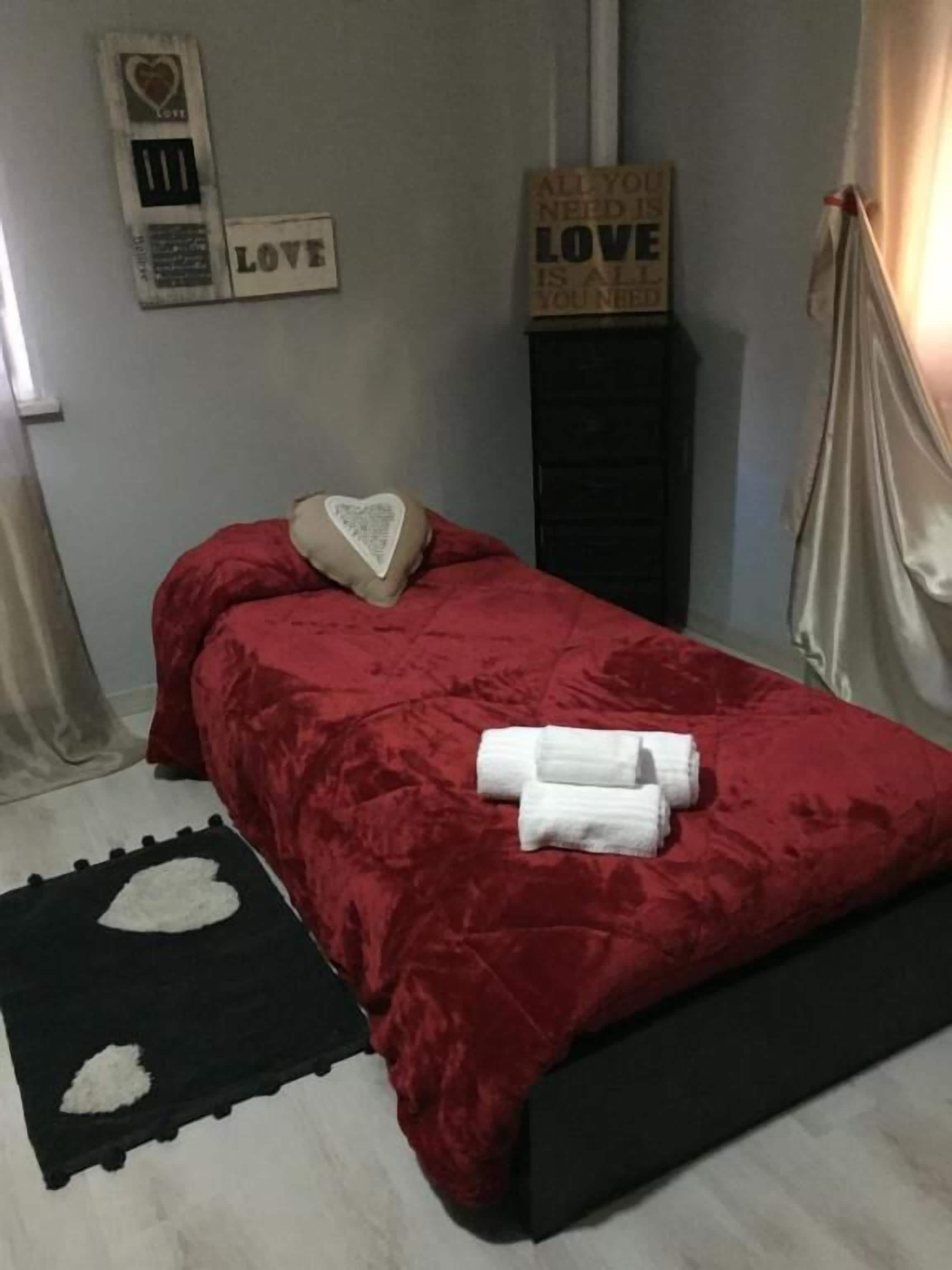 A neatly made bed with a red bedspread and a heart-shaped pillow, a black and white rug, and a sign on the wall that says "ALL YOU NEED IS LOVE".