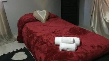 A neatly made bed with a red bedspread and a heart-shaped pillow, a black and white rug, and a sign on the wall that says "ALL YOU NEED IS LOVE".