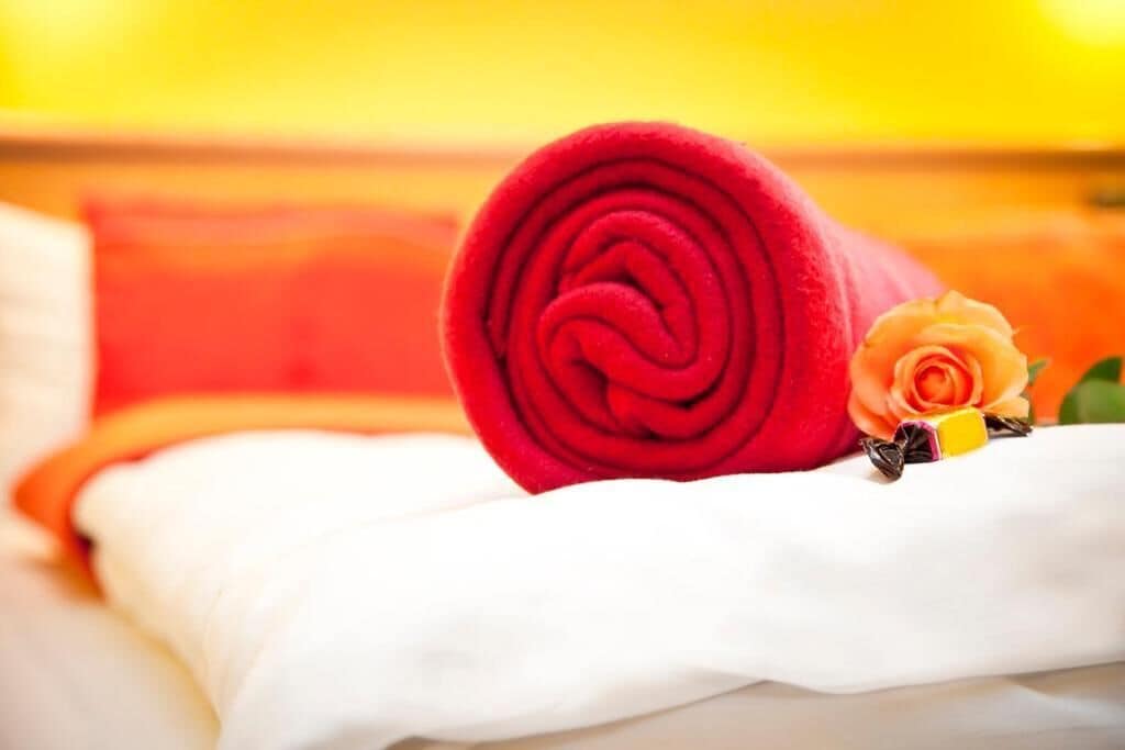 Una toalla roja enrollada con cuidado sobre una cama, junto a un almohadón blanco y una rosa amarilla.