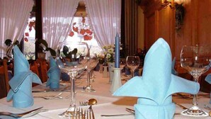 A dining table set with blue napkins, silverware, and wine glasses, with a floral arrangement in the background.