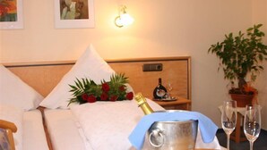 A hotel room with a bed, a bedside table with a bottle and glasses, a potted plant, and a wall-mounted light.