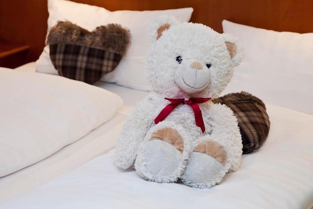Ein weiches, weißes Kuscheltier mit roter Schleife auf einem Bett mit weißer Bettwäsche und karierten Kissen.