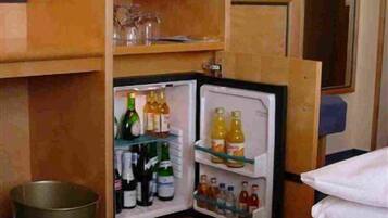 A compact hotel room with a small television on a wooden stand, a mini-fridge stocked with beverages, and a trash bin.