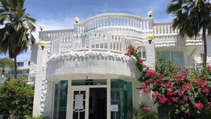 Un bâtiment blanc doté d’un balcon incurvé et d’une entrée en verre. Des plantes et des fleurs en pot sont disposées autour de l’entrée.