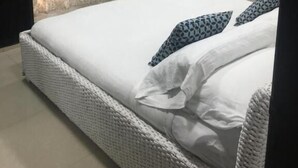 Un lit bien fait, avec du linge de lit blanc et des coussins à motifs, une table de chevet ornée d’un vase de fleurs et un rideau à motifs.