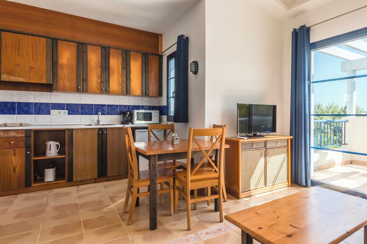 Cocina moderna con armarios de madera, comedor con mesa y sillas, y balcón con mobiliario exterior.