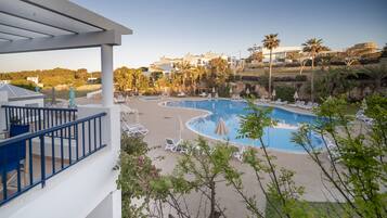 A pool area with lounge chairs and umbrellas, surrounded by greenery and buildings in the background.