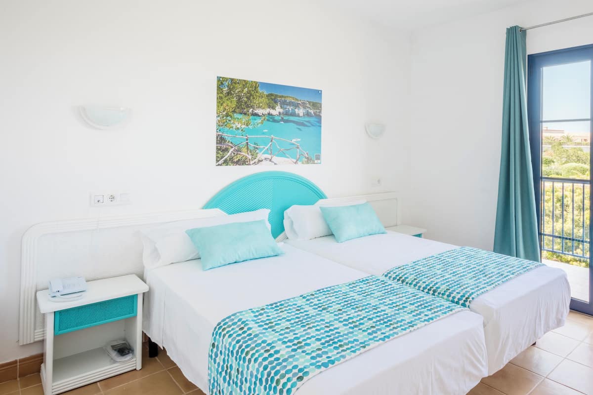 Habitación de hotel con cama, mesita de noche, ventana con vistas y puerta a un balcón exterior.