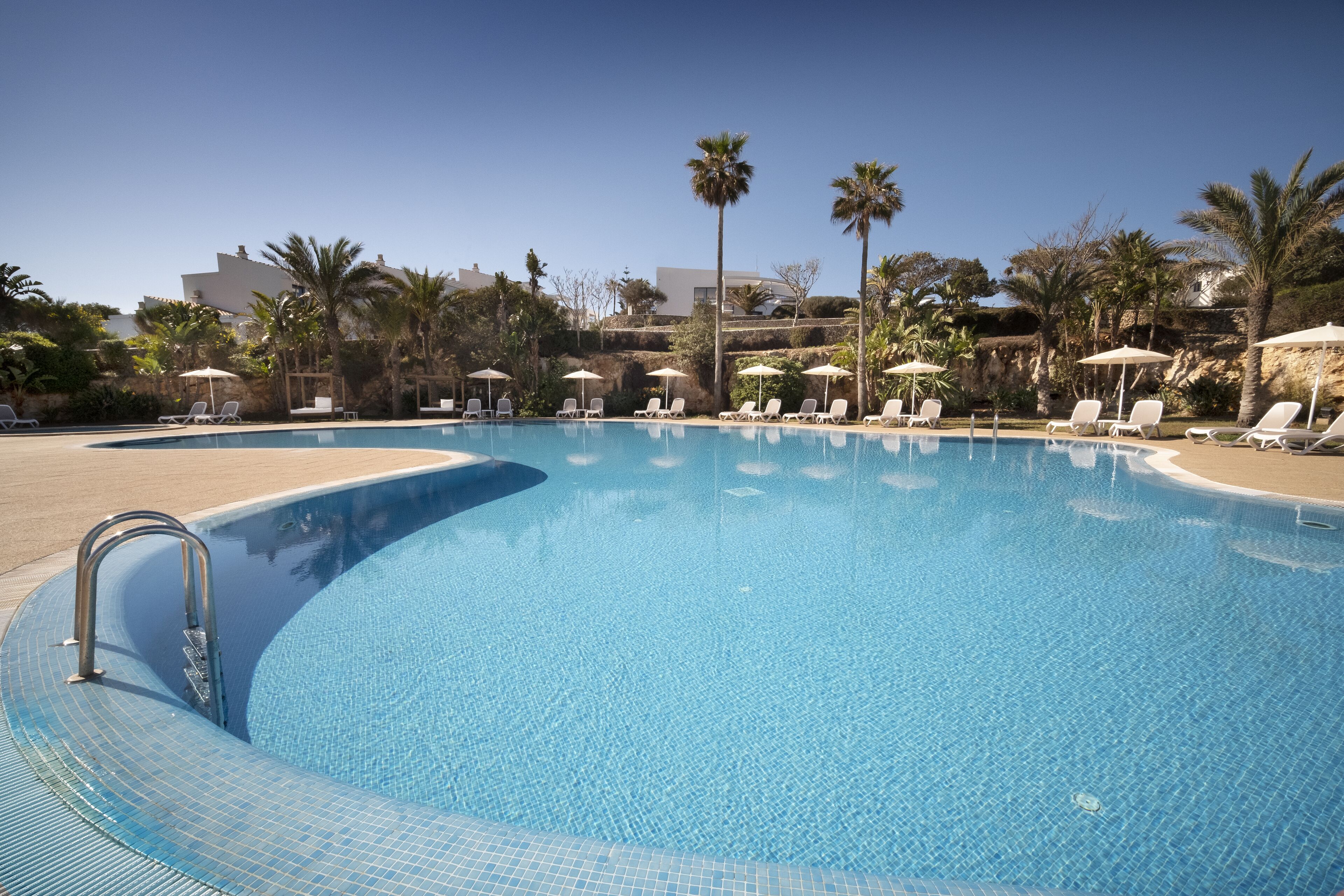 A clear blue swimming pool with lounge chairs and umbrellas, surrounded by palm trees and buildings.