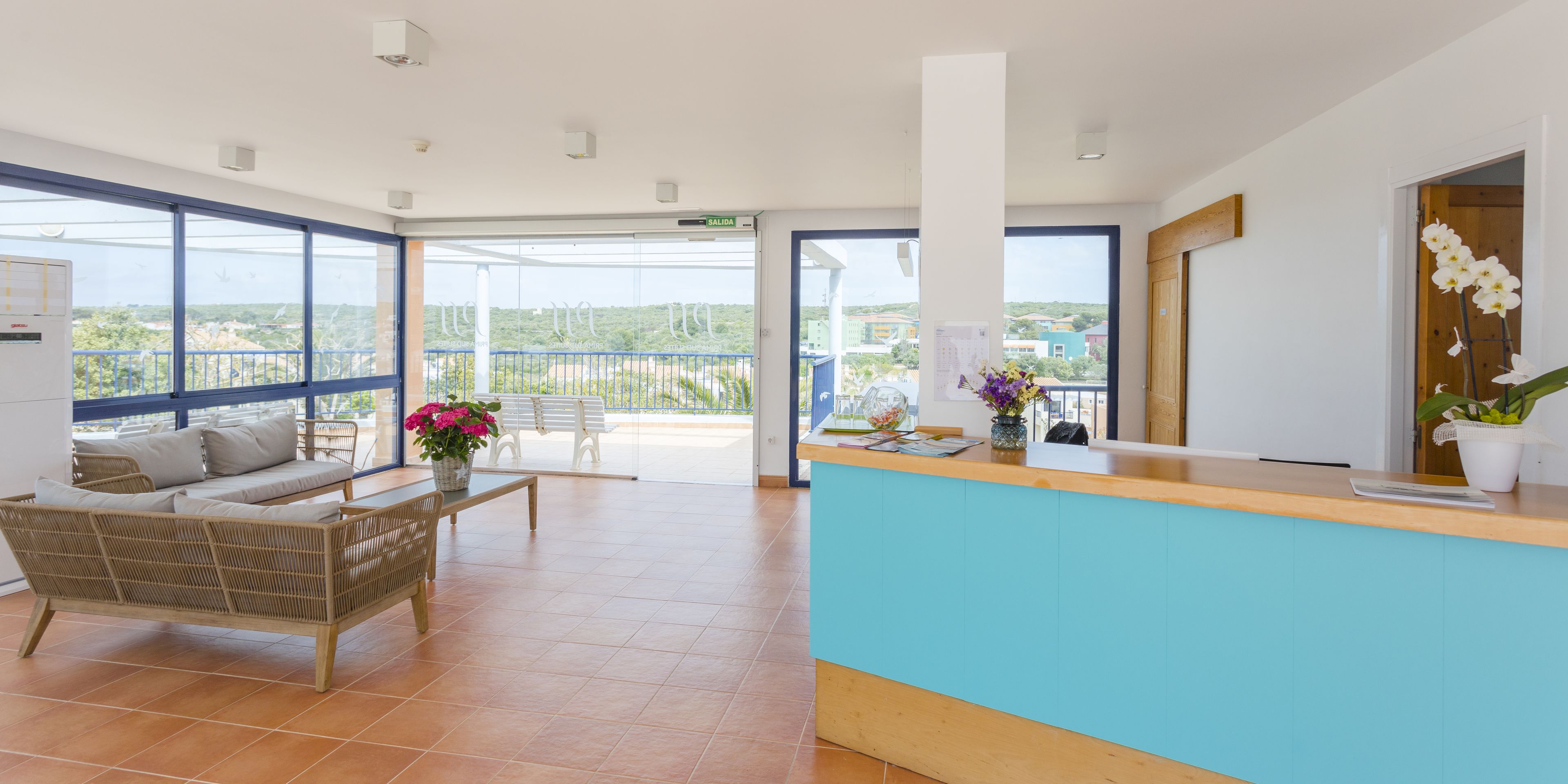 A spacious reception area with a blue counter, a wicker sofa, a wooden bench, and a view of the city through large windows.