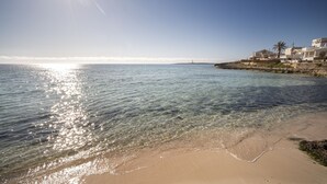 A beach with clear water, a sandy shore, and buildings in the background. -  Bimbolla Apartaments (Sant Lluis)