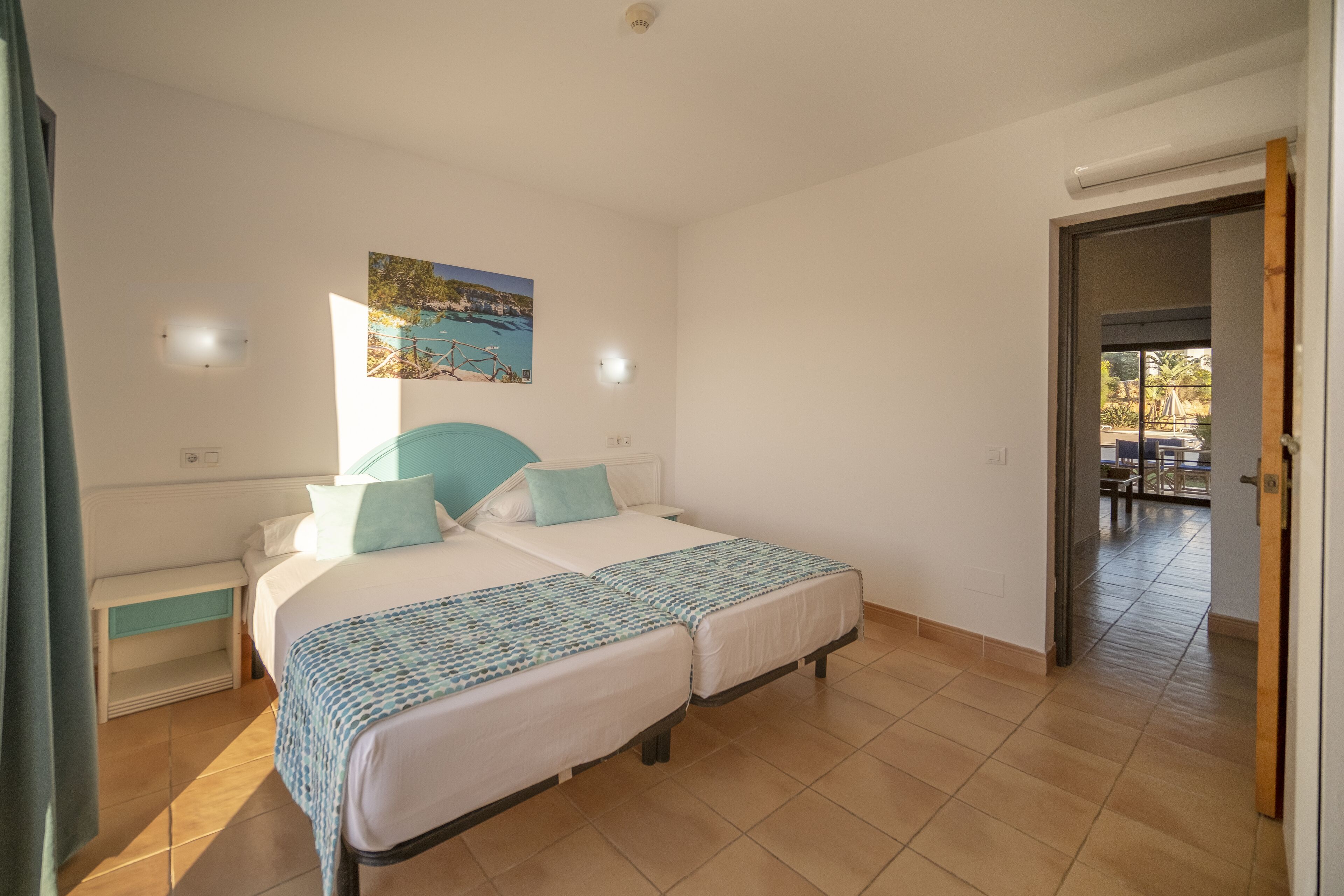 A bedroom with two beds, a painting on the wall, and a view of the outside balcony.
