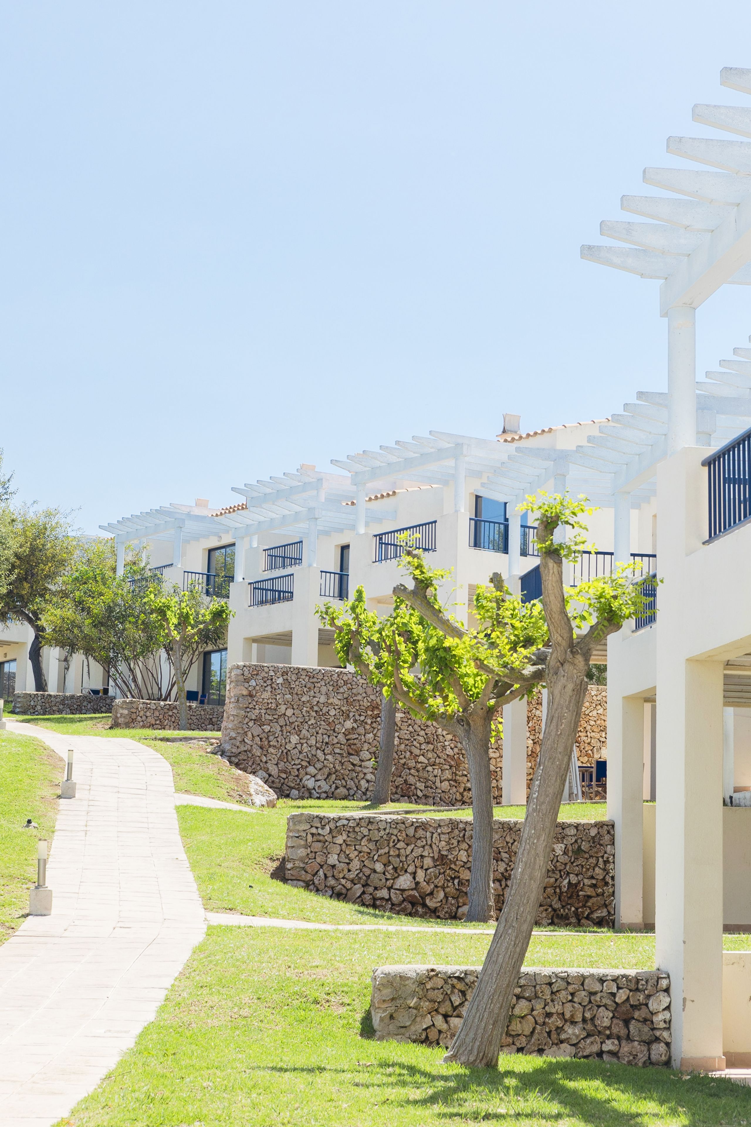 A pathway leading to a row of white buildings with balconies, stone walls, and trees.