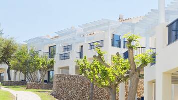 A pathway leading to a row of white buildings with balconies, stone walls, and trees.