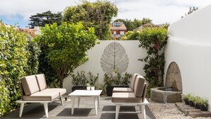 En udendørs terrasse med to beige sofaer, et hvidt bord og et dekorativt bladmaleri på væggen.