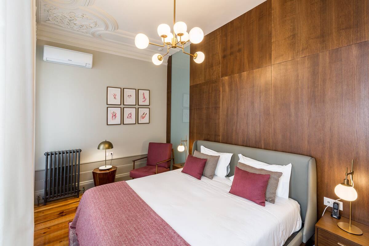 A modern hotel room with a large bed, a wooden floor, a radiator, a wall-mounted air conditioner, and a chandelier.