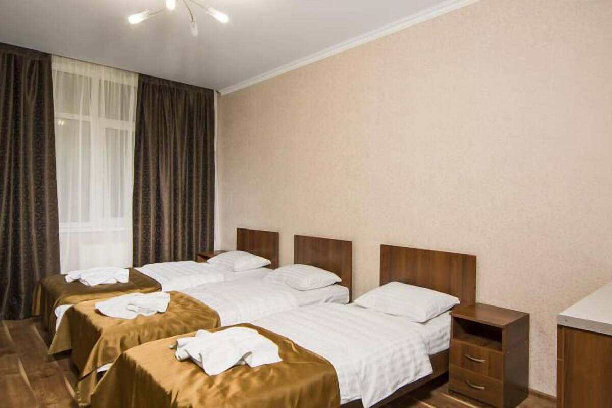 A hotel room with two beds, a wooden headboard, a bedside table with drawers, and a large window with curtains.