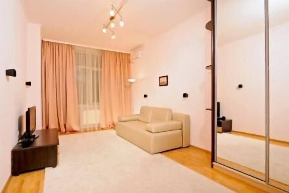 A modern living room with a beige sofa, a flat-screen TV on a wooden stand, and a large mirror on the wall.