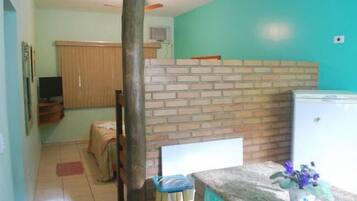 Quarto pequeno e compacto, com divisória de tijolos, uma cama, uma escrivaninha e um frigorífico.