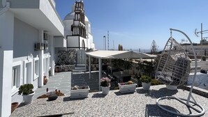 A courtyard with a white building, a bell tower, potted plants, outdoor seating, and a metal sculpture. - Villa Pavlina (Santorini)