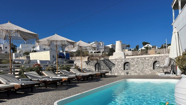 A pool area with lounge chairs and umbrellas, surrounded by stone walls and white buildings. - Villa Pavlina (Santorini)