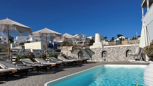 Zona de piscina con sillones de descanso y sombrillas, rodeada de muros de piedra y edificios blancos.