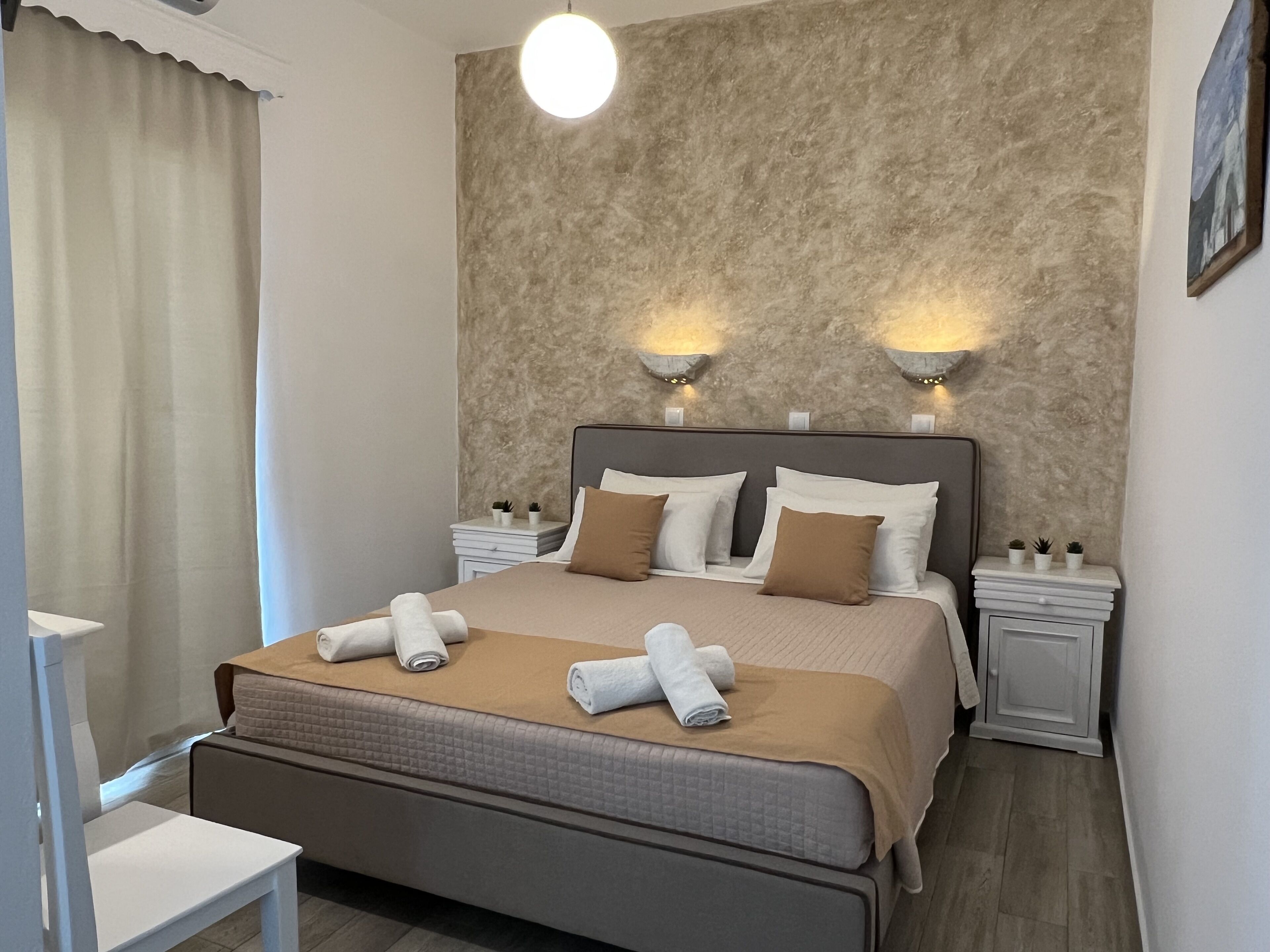 A neatly arranged hotel room with a large bed, two bedside tables, a nightstand, a wall-mounted light fixture, and a window with curtains.
