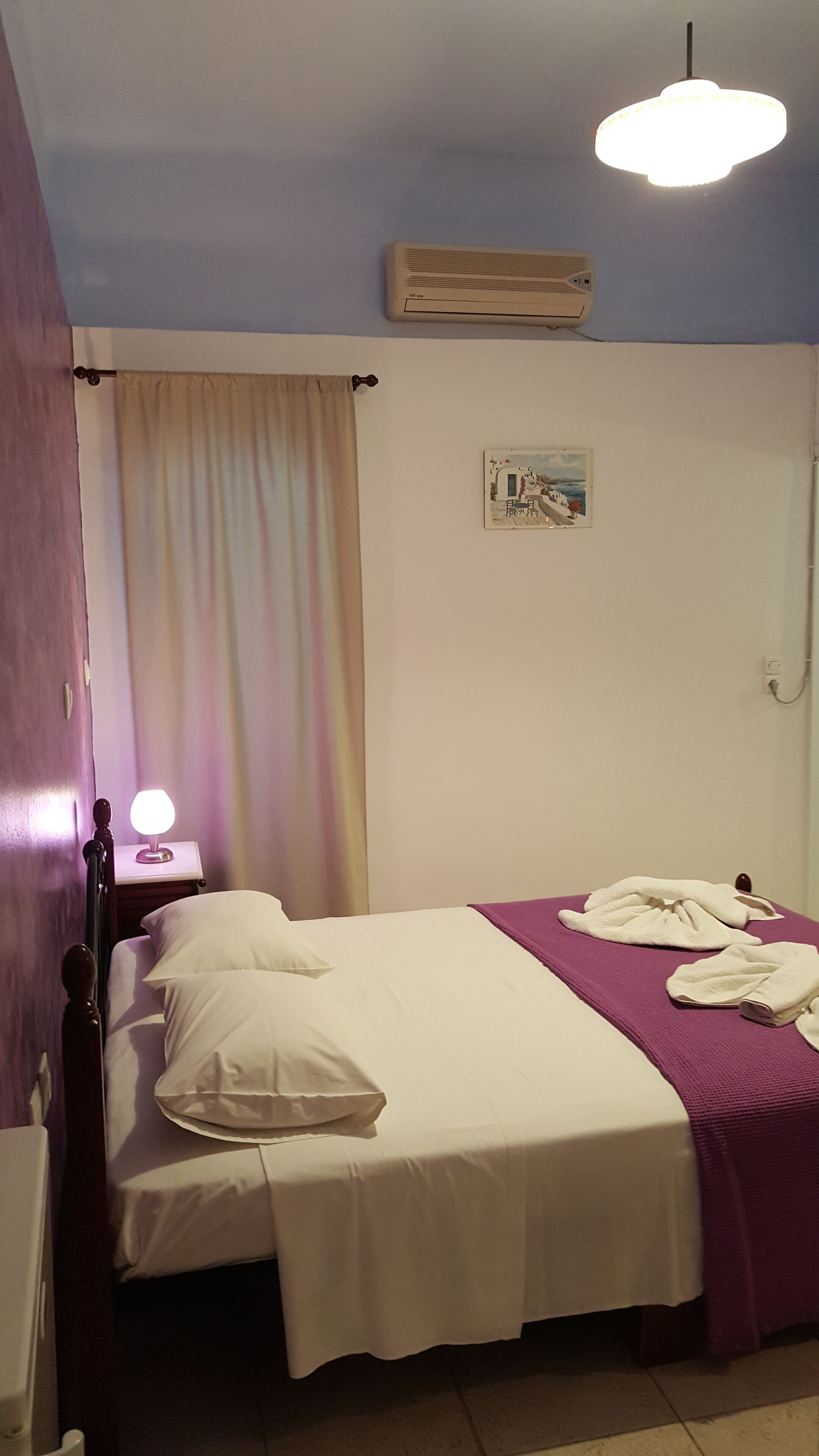 A bedroom with a bed, a nightstand, a lamp, a painting, and an air conditioner.
