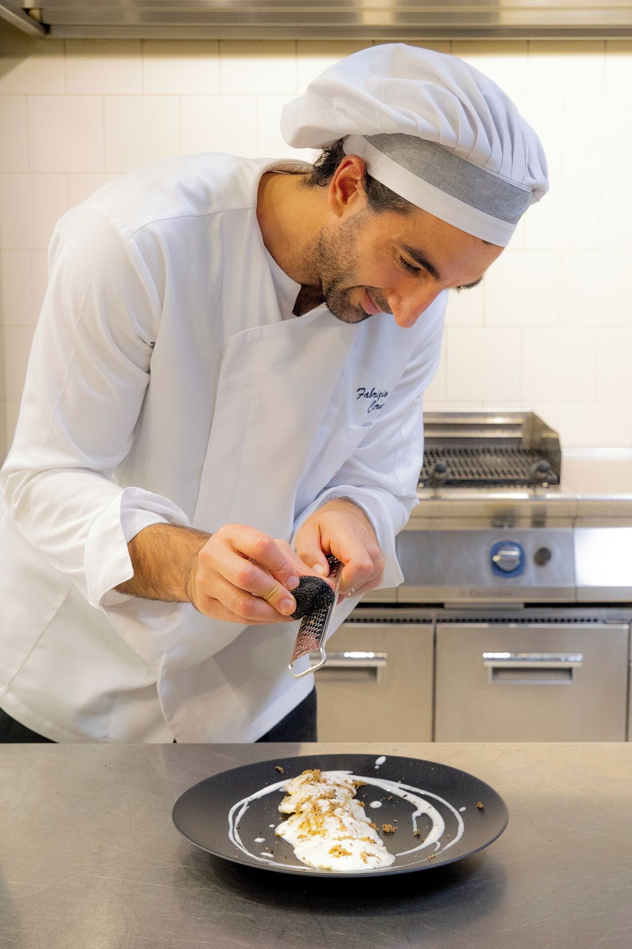 A chef in a white uniform and hat, grating cheese on a black plate in a kitchen.