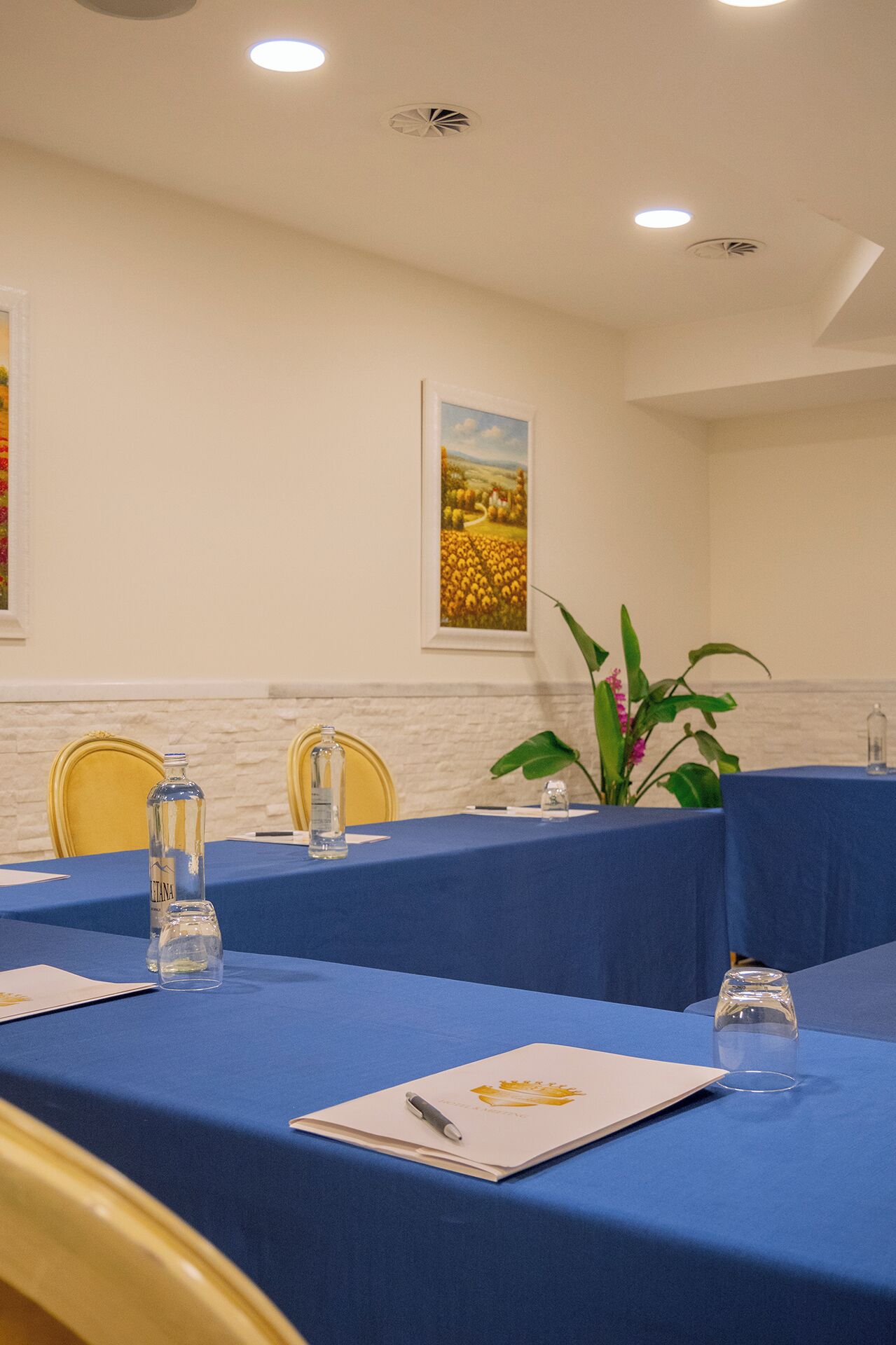 Sala de conferência com toalhas de mesa azuis, cadeiras, garrafa de água, copo, caneta e um quadro na parede.