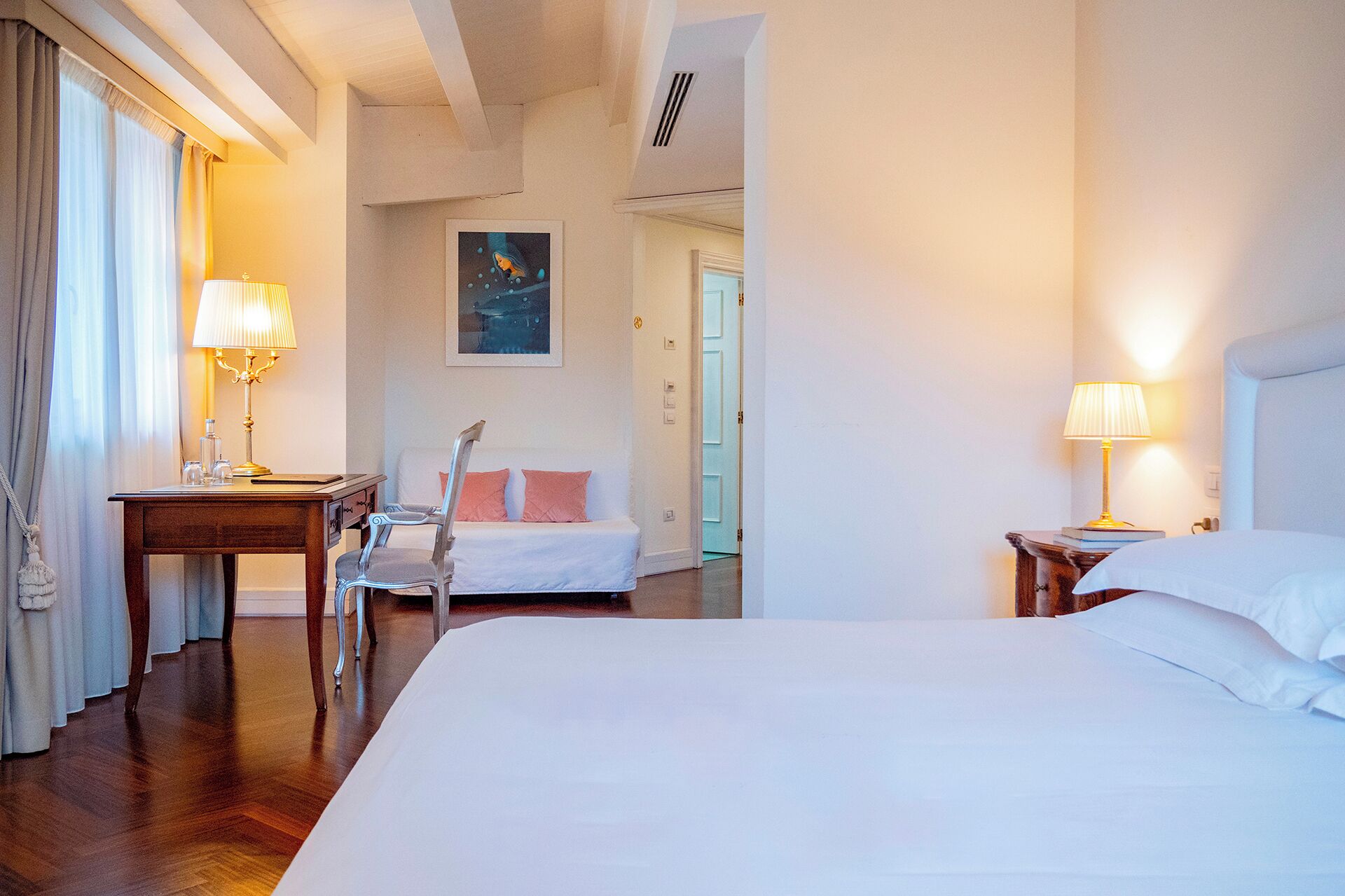 A hotel room with a large bed, two bedside tables with lamps, a chair, a desk with a lamp, and a painting on the wall.