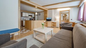 A modern living room with a sofa, a coffee table, and a TV. There is a kitchen in the background with wooden cabinets and a dining area. - Landhaus Edelweiss (Sankt Leonhard im Pitztal)
