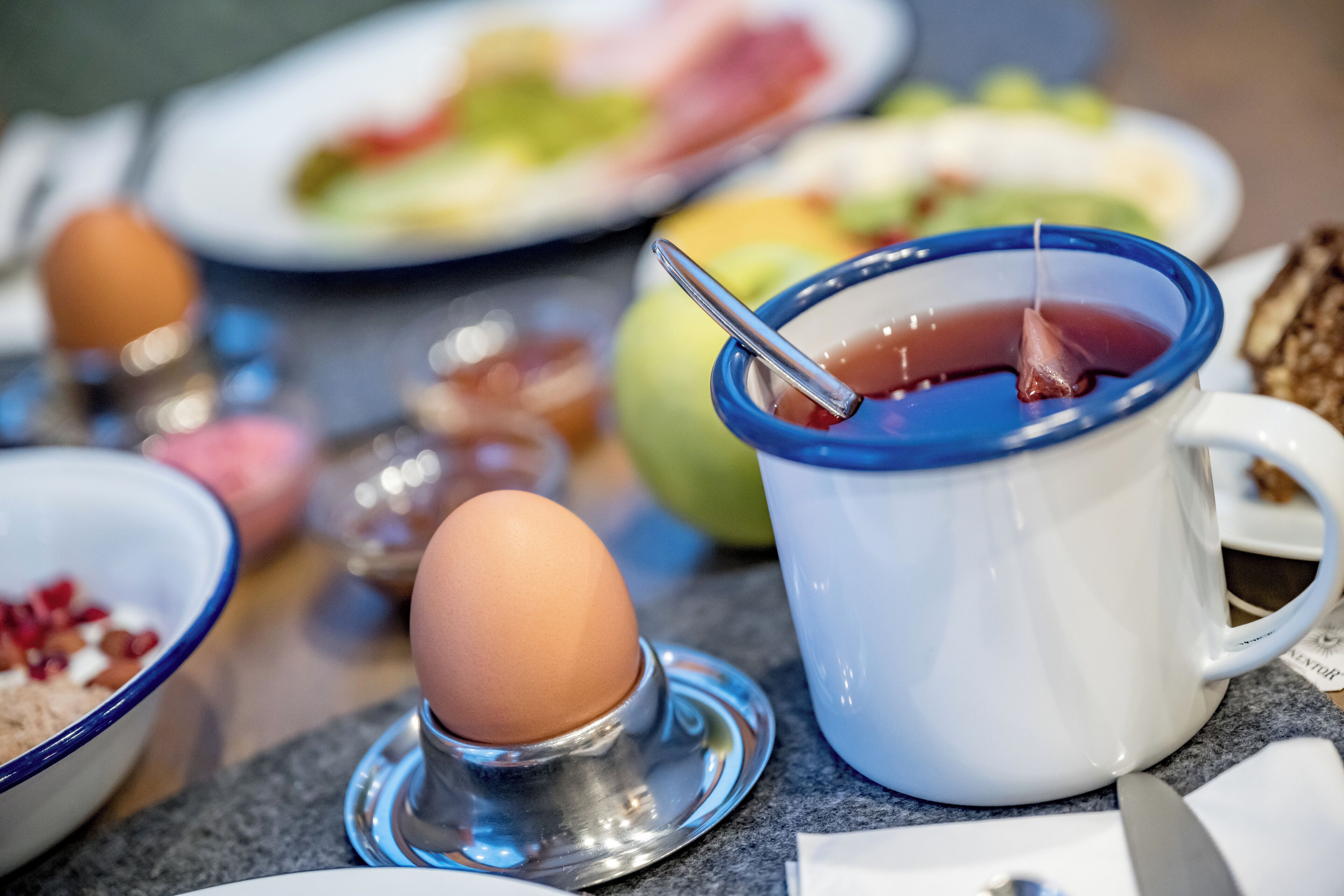 Una mesa con una taza azul que contiene líquido rojo, un soporte metálico para huevos con un huevo marrón, y varios platos al fondo.