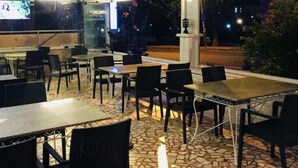 Un área de comedor al aire libre con mesas y sillas, piso de baldosas y una pantalla de televisión mostrando un programa.