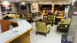 Un vestíbulo de hotel con mostrador de recepción, asientos tapizados en verde y una zona de comedor con mesas y sillas.
