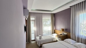 Habitación de hotel con dos camas, una mesita de noche de madera, una lámpara y vistas al exterior a través de amplias ventanas.