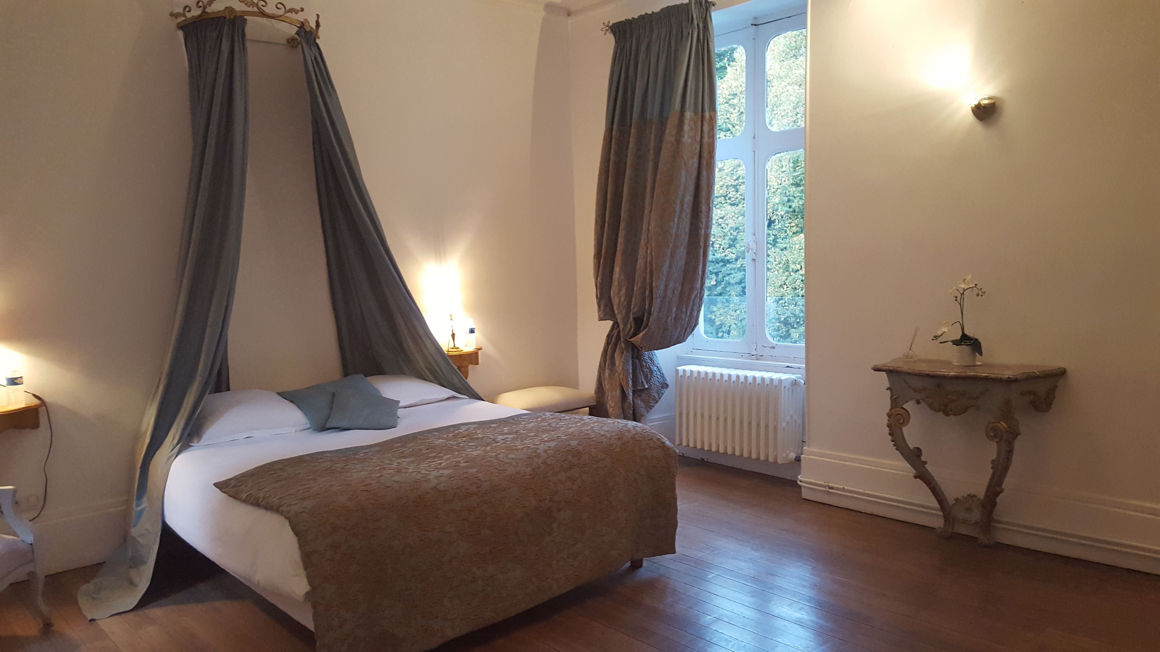 A bedroom with a four-poster bed, a bedside table with a lamp, a radiator, and a window with curtains.