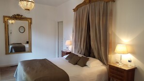 A hotel room with a large bed, a mirror with an ornate frame, a chandelier, a bedside table with a lamp, and a curtain.