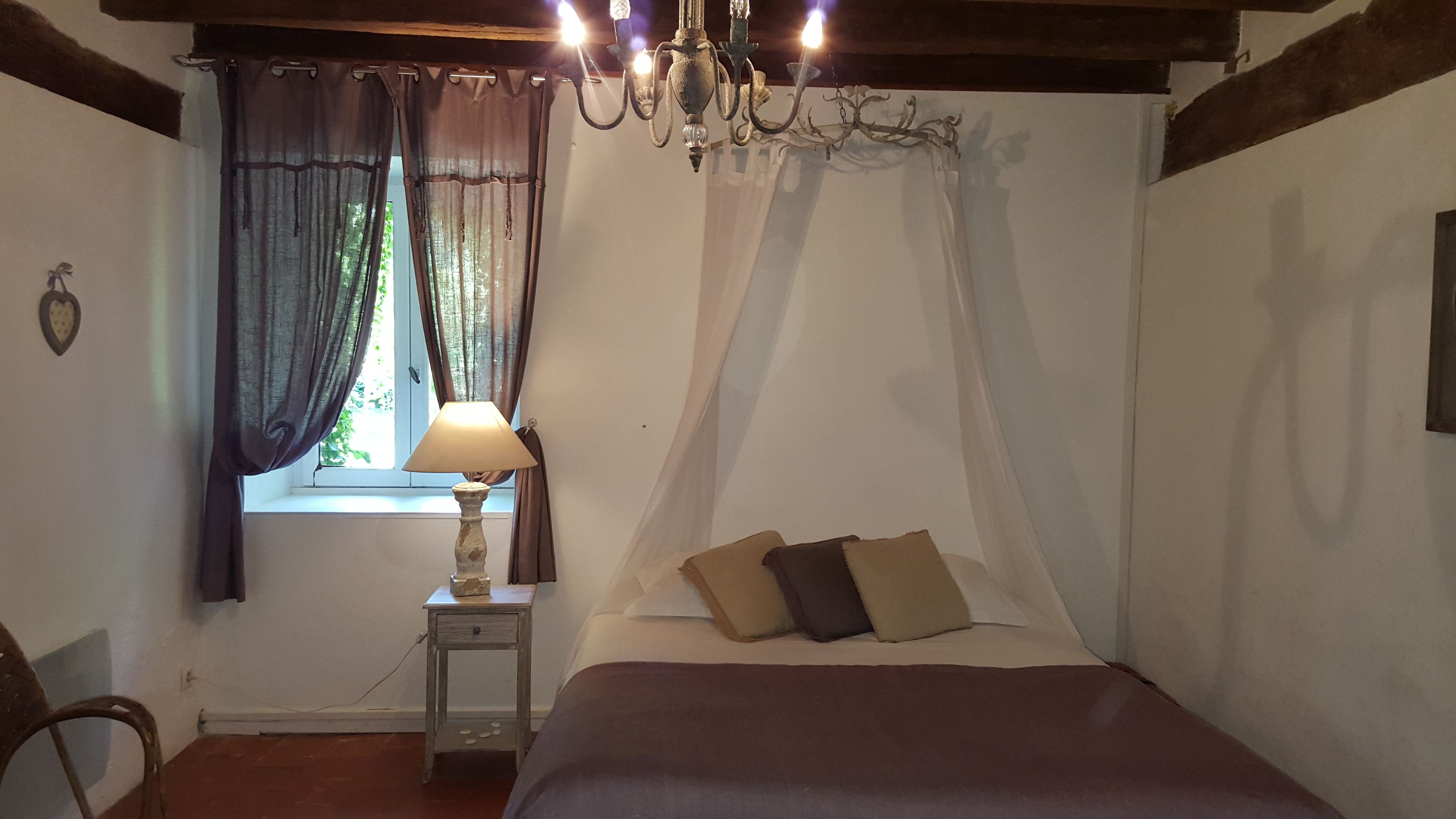 A bedroom with a bed, a nightstand with a lamp, a window with curtains, and a chandelier.