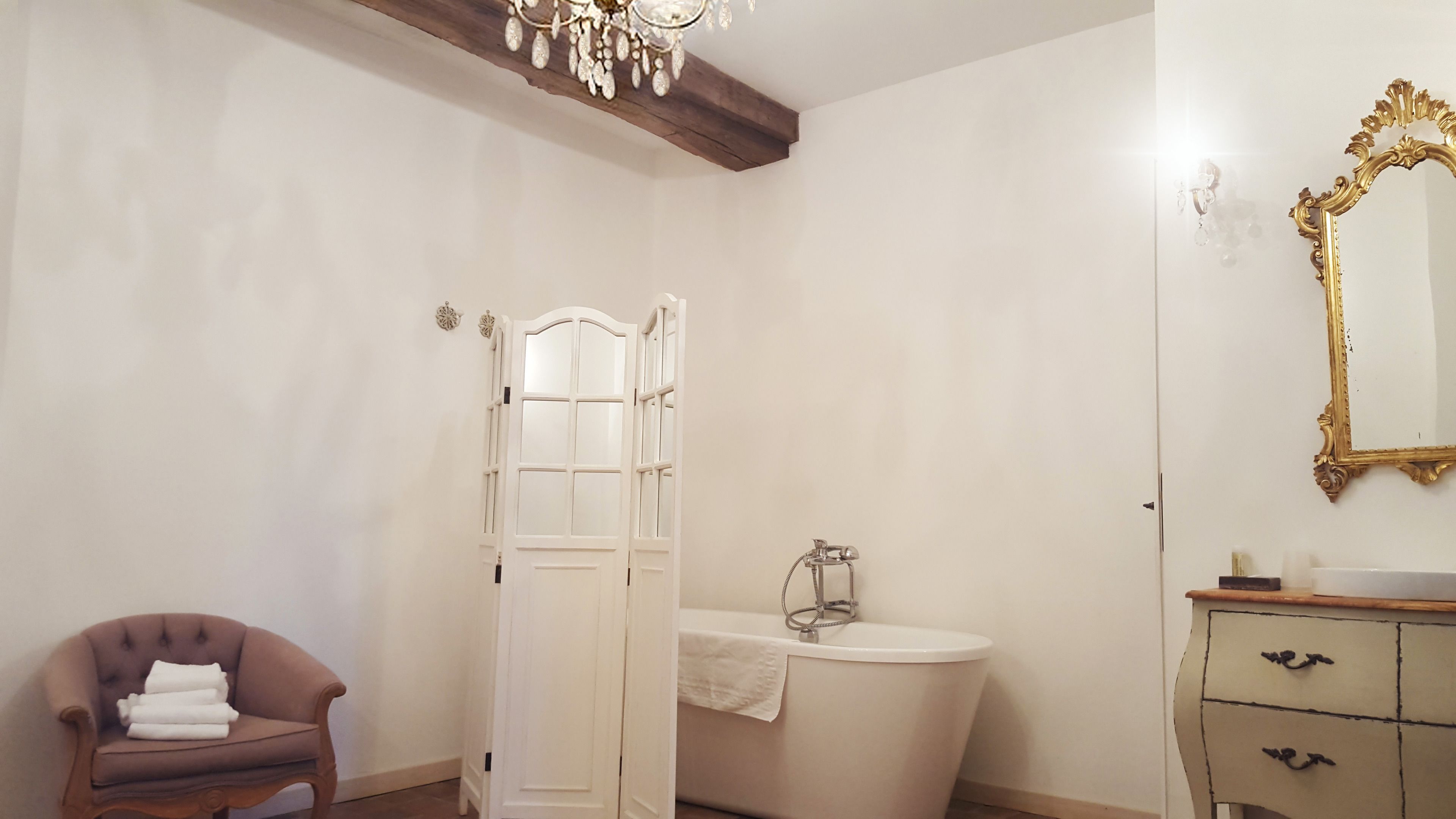 Un baño con una bañera blanca, un tocador con espejo de marco dorado, un sillón marrón con una toalla y una viga de techo de madera.