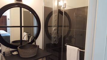 Une salle de bain avec un miroir rond, un lavabo surmonté d’un récipient, un porte-serviettes et une douche dotée d’une porte en verre.
