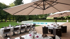 Un área de comedor al aire libre con mesas dispuestas para una comida, rodeada de sombrillas y con vistas a una piscina.